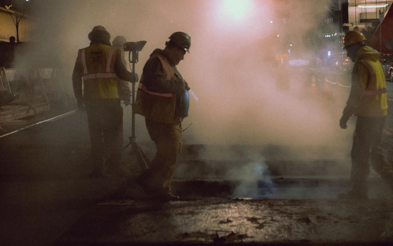 Road crew in high-visibility vests working around a steam vent on a city street at night
